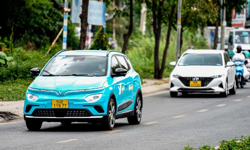 Hình ảnh một chiếc taxi điện VinFast màu xanh đang hoạt động trên đường phố Hà Nội nhộn nhịp
